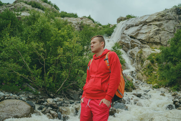 Portrait of young man looking away on background of waterfall. Adult male tourist enjoying beautiful view in mountainous area. - Fotografie, Obrázek