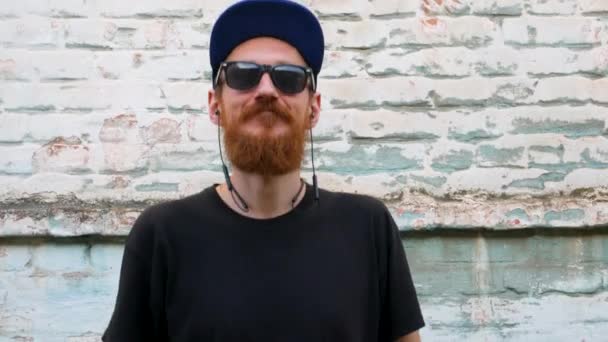 Portrait of young red bearded man in black t-shirt and snapback in rock style on urban background listening to music with headphones. Hipster guy in wireless earphones on old blue brick wall backdrop. - Footage, Video