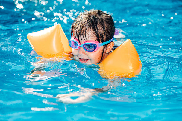 Маленька чорноволоса дівчинка посміхається і плаває в басейні в рукавах і окулярах для плавання
 - Фото, зображення