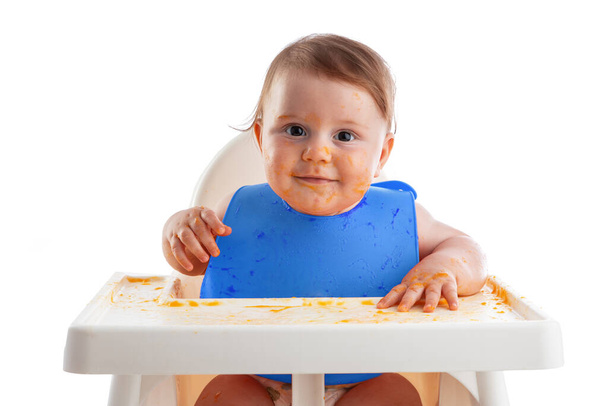 Fröhliches Kleinkind isst Lebensmittel selbst mit den Händen. Porträt eines glücklichen, schmutzigen Jungen im Hochstuhl und chaotisch herum. - Foto, Bild
