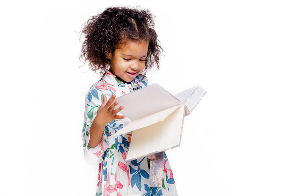 Kleine hübsche afrikanisch-amerikanische Mädchen in weißem Blumenkleid schauen auf Notizbuch - Foto, Bild