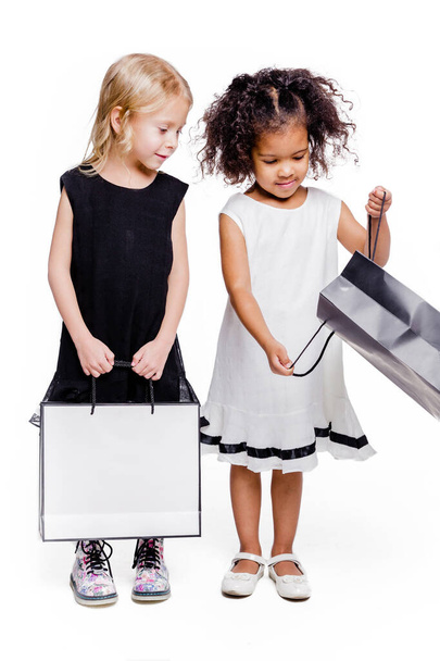 Little fashionable girls of two different nationalities are posing against a white background with bags from the store. One girl shows the other what is in her bag - Fotó, kép