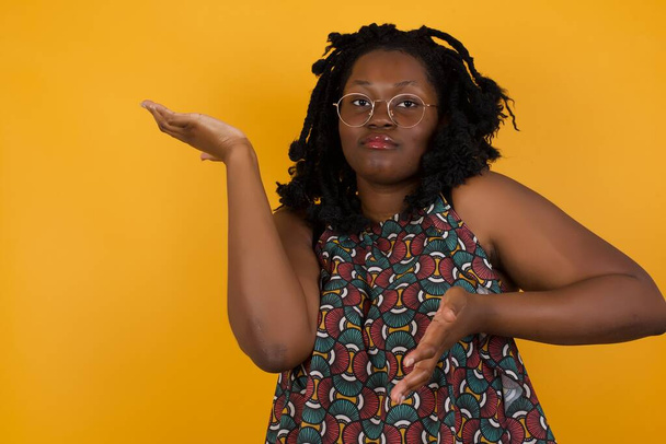 Jonge mooie Afro-Amerikaanse vrouw met een bril over gele achtergrond die met beide handen opzij wijst en iets vreemds laat zien en zegt: Ik weet niet wat dit is. Reclameconcept. - Foto, afbeelding