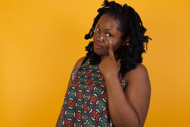 Jovem mulher afro-americana bonita usando óculos sobre fundo amarelo Apontando para o olho observando você gesto, expressão suspeita
.  - Foto, Imagem