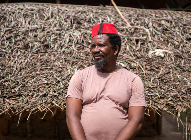 Un homme plus âgé africain en rouge musulman Taqiyyah Fez Chapeau posant avec un bâton pour les personnes boiteuses sur la cour près de la cabane de base avec toit de chaume dans un petit village isolé en Tanzanie, île de Pemba, Zanzibar - Photo, image