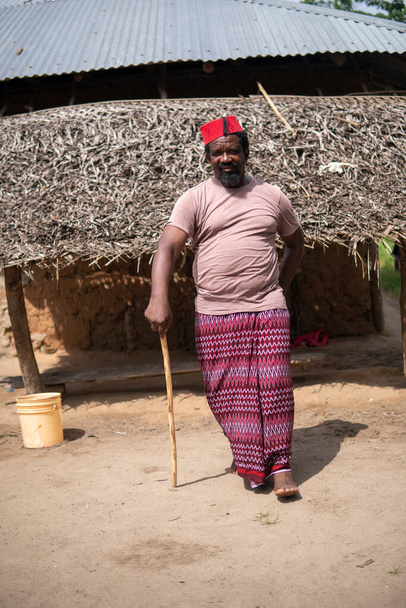 Un hombre mayor africano en rojo musulmán Taqiyyah sombrero de Fez posando con un palo para las personas cojas en el patio cerca de la cabaña básica con techo de paja en la pequeña aldea remota en Tanzania, isla de Pemba, Zanzíbar - Foto, Imagen