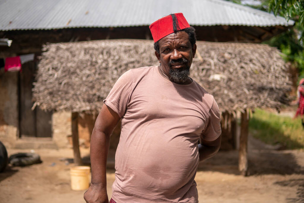 Een Afrikaanse oudere man in rode moslim Taqiyyah Fez Hoed poseren met een stok voor lamme mensen op Yard In de buurt van de Basic Hut met Thatched dak in Small Remote Village in Tanzania, Pemba eiland, Zanzibar - Foto, afbeelding