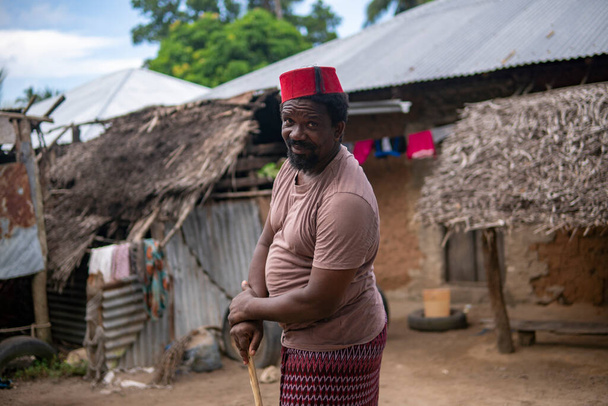 Kızıl Müslüman Takiyyah Fez Şapkalı Afrikalı yaşlı bir adam, Tanzanya 'nın Küçük Uzak Köyü' nde, Pemba Adası 'nda, Zanzibar' da çatısı Thatched olan, Yard 'da topal insanlar için sopayla poz veriyor. - Fotoğraf, Görsel