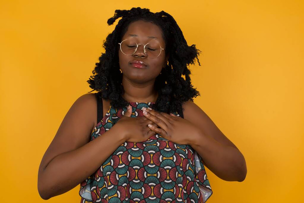 Young beautiful afro american woman wearing glasses over yellow background  smiling with hands on chest with closed eyes and grateful gesture on face. Health concept. - Фото, изображение