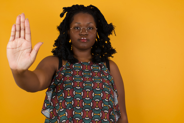 Mladá krásná afro americká žena s brýlemi přes žluté pozadí dělá stop gesto dlaní ruky. Varovný výraz s negativním a vážným gestem na tváři  - Fotografie, Obrázek
