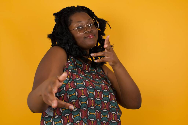 Jovem mulher afro-americana bonita vestindo óculos sobre fundo amarelo piscando e apontando com pistolas de dedo para a câmera feliz e insolente posando. Ei, você, bang
. - Foto, Imagem