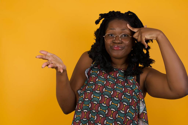 Jovem mulher afro-americana bonita vestindo óculos sobre fundo amarelo confuso e irritado com a palma aberta mostrando espaço de cópia e apontando o dedo para a testa. Pense nisso.
. - Foto, Imagem