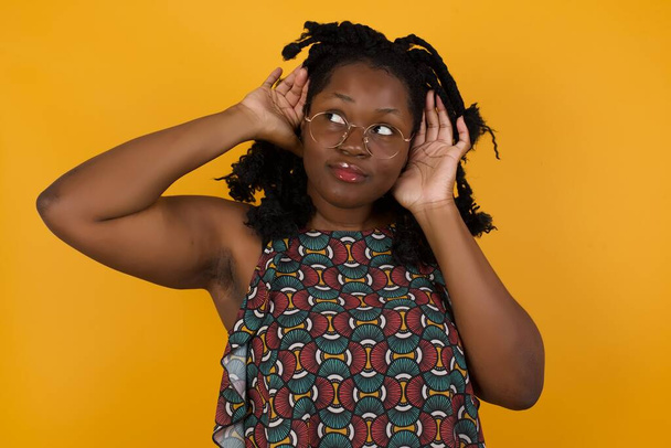 Молодая красивая афро-американка в очках на жёлтом фоне пытается услышать жест обеих рук на ухе, любопытный для сплетен. Проблемы со слухом, глухой - Фото, изображение