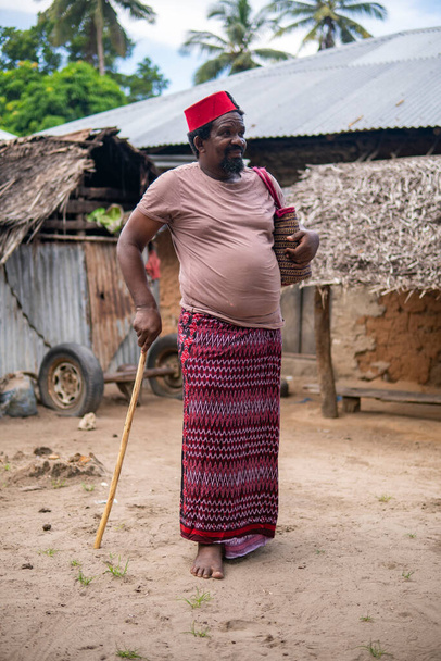 Un uomo anziano africano in rosso musulmano Taqiyyah Fez cappello in posa con un bastone per le persone zoppe su Yard Near the Basic Hut con tetto Thatched in Small Remote Village in Tanzania, isola di Pemba, Zanzibar - Foto, immagini