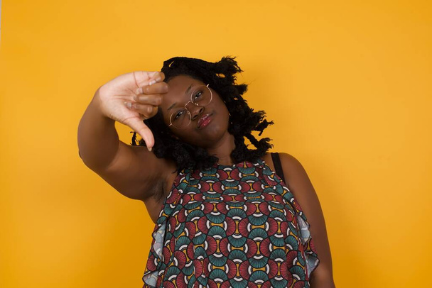 Young beautiful afro american woman wearing glasses over yellow background feeling angry, annoyed, disappointed or displeased, showing thumbs down with a serious look  - 写真・画像