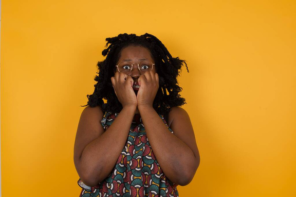 Fotografie ustrašené Mladá krásná afro americká žena nosí brýle přes žluté pozadí drží ruce u úst, cítí strach a strach, má fóbii, Má Afro účes. Šok a zděšený koncept. - Fotografie, Obrázek