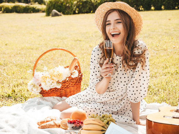 Young beautiful hipster woman in trendy summer sundress and hat. Carefree woman making picnic outside. Positive model sitting on plaid on grass, eating fruits and cheese, drinking champagne - Foto, Imagen