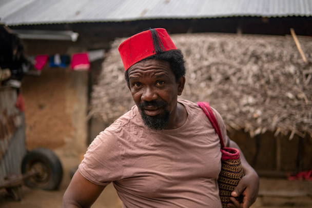 Portré egy afrikai idősebb férfi a piros muszlim Taqiyyah Fez kalap pózol egy bottal a sánta emberek a Yard közelében az alapvető kunyhó Thatched tető Small Remote Village Tanzánia, Pemba sziget - Fotó, kép