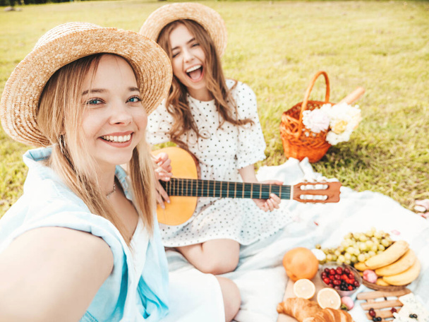 Two young beautiful hipster woman in trendy summer sundress and hats. Carefree women making picnic outside. Positive models sitting on plaid on grass, eating fruits and cheese. Playing the guitar - Valokuva, kuva