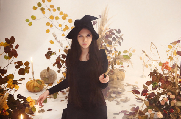 Jonge vrouw in heks kostuum kijken naar de camera en het gebruik van toverstok, terwijl staan op de achtergrond van droge bloemige compositie tijdens Halloween viering - Foto, afbeelding