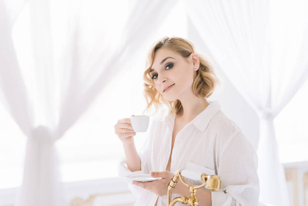 Hermosa mujer rubia joven en una camisa blanca en un baño ligero con plomería de oro está disfrutando de una taza de café por la mañana. Enfoque selectivo suave. - Foto, imagen