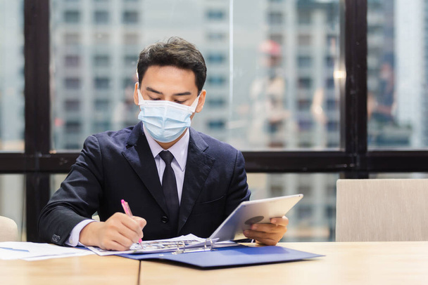 asian young businessman wear protective mask using digital tablet and writing data on paper work in office. man wear mask protection during spread of coronavirus or covid-19. - 写真・画像