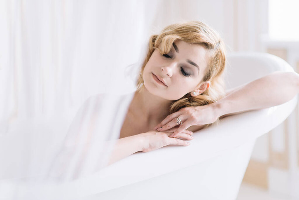 Joven hermosa chica se relaja en un baño blanco con accesorios de oro en una habitación luminosa. - Foto, imagen