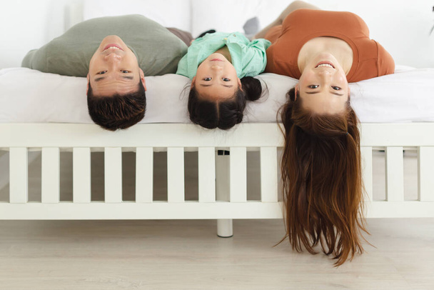szczęśliwy Azji rodzina relaks w wakacje w sypialni w domu, ojciec matka i córka leżące do góry nogami wisząca głowa na łóżku. - Zdjęcie, obraz