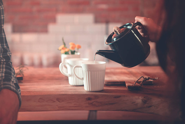 Kobieta Crop z czajniczek wlanie herbaty do filiżanki podczas siedzenia z mężem przy drewnianym stole w kuchni domowej - Zdjęcie, obraz