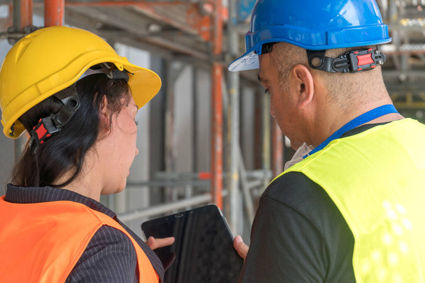 Deux travailleurs de la construction dos tourné portant un gilet de sécurité, un casque et des lunettes vérifiant leurs plans de travail sur une tablette numérique - Photo, image
