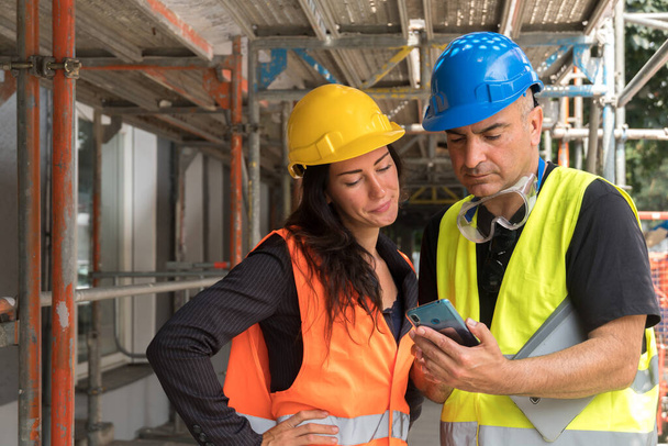 Deux travailleurs sur le chantier portant un gilet de sécurité, un casque et des lunettes vérifiant le plan de travail sur une tablette numérique - Photo, image