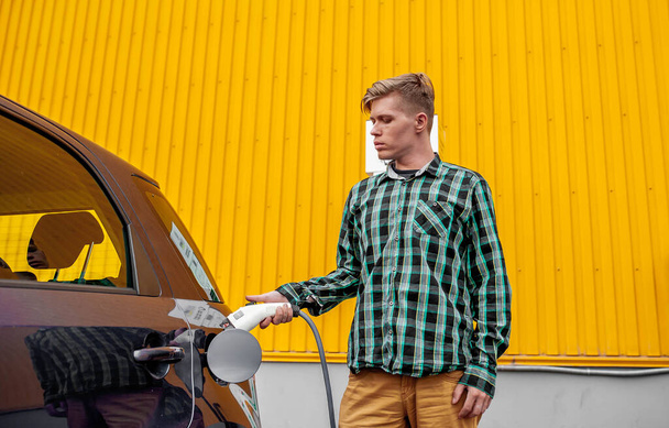 młody przystojny biały mężczyzna stoi na parking i ładuje jego elektrosamochód - Zdjęcie, obraz