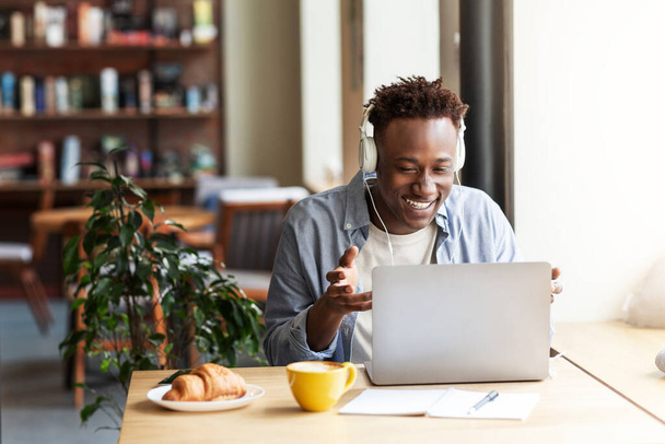 Glücklicher schwarzer Kerl mit Kopfhörern kommuniziert online vom Laptop-Computer im Café, Kopierraum - Foto, Bild