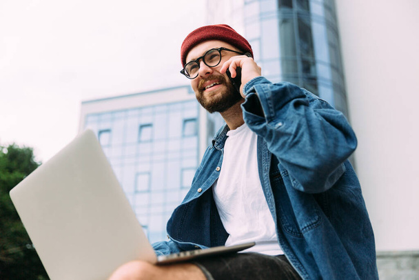 Portret współczesnego brodatego hipstera w okularach rozmawiającego przez telefon i korzystającego jednocześnie z laptopa. - Zdjęcie, obraz