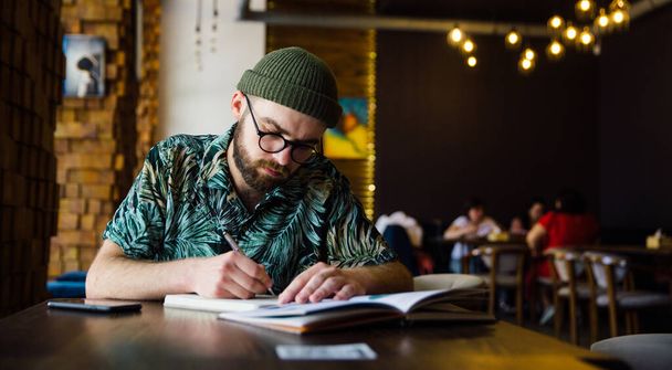 Bearded oldschool young handsome romantic writer writing poem in notebook in cafe. Room for text. - Foto, Imagen