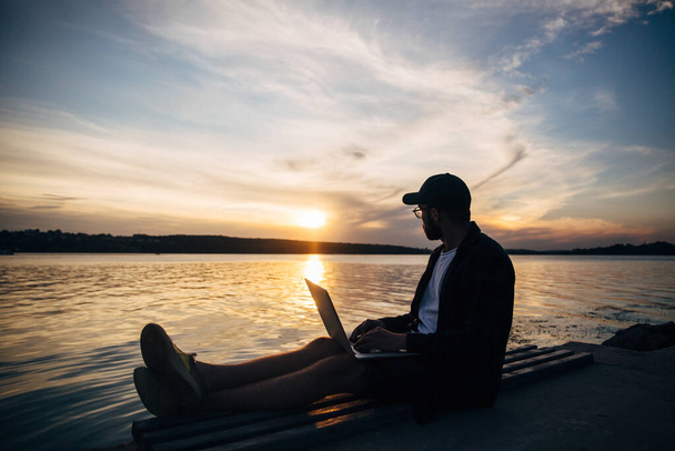 Silhouette of freelancer hipster male in baseball cap working on laptop on riverside in park and enjoying sunset. Room for text. - Foto, immagini