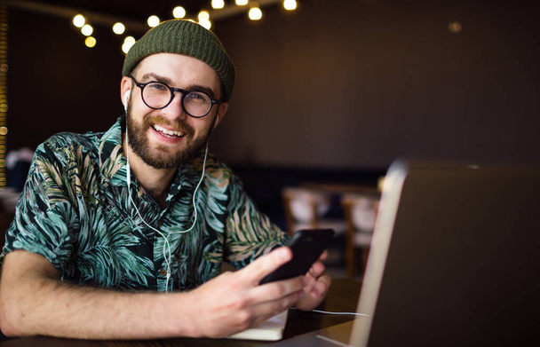 Šťastný veselý módní mužský hipster ve sluchátkách pomocí notebooku a smartphonu dívající se do kamery a usmívající se. Technologický koncept. - Fotografie, Obrázek