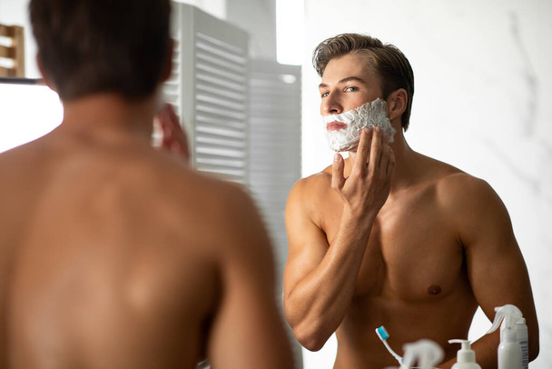 Handsome young man looking in the mirror applying foam - Photo, Image