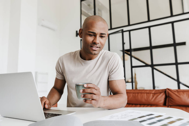 Fotografie potěšeného africký Američan muž pracuje s notebookem a pití kávy u stolu v obývacím pokoji - Fotografie, Obrázek