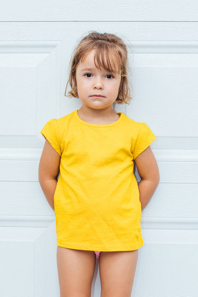 Adorável menina de cabelos loiros vestindo uma camisa amarela encostada ao fundo branco
 - Foto, Imagem