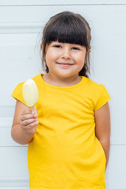 黒髪のかわいい女の子はアイスクリームを食べ、白い背景にもたれて黄色のシャツを着て - 写真・画像