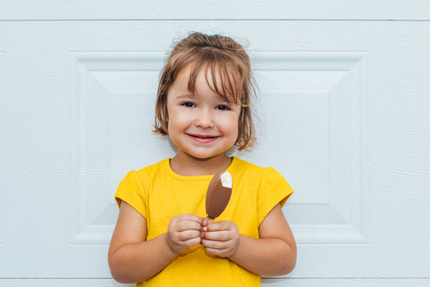 Adorabile ragazza dai capelli biondi che mangia gelato, indossando una camicia gialla appoggiata sullo sfondo bianco - Foto, immagini