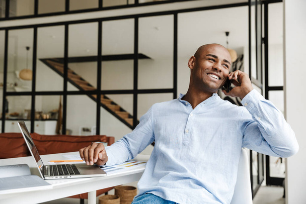Фото радостного африканского американца, разговаривающего по мобильному телефону во время работы с ноутбуком в гостиной - Фото, изображение