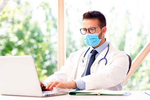 Portrait of male doctor wearing face mask and working on laptop. - Fotó, kép