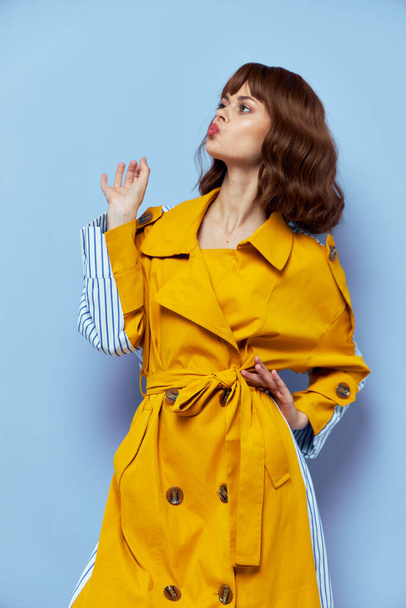 Een brunette in een gele jas met een witte doek stuurt een luchtkus op een blauwe achtergrond - Foto, afbeelding