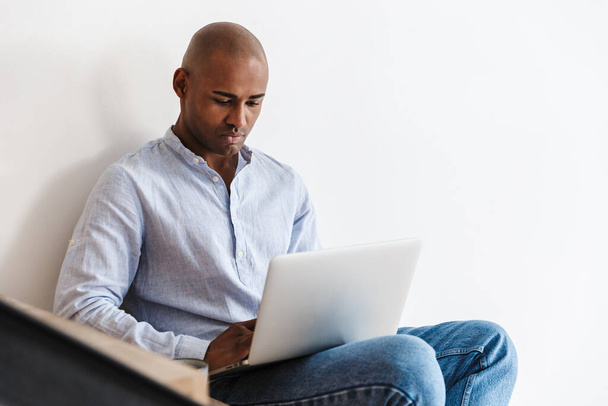 Photo of focused african american man working with laptop while sitting on wooden stair at home - Photo, image