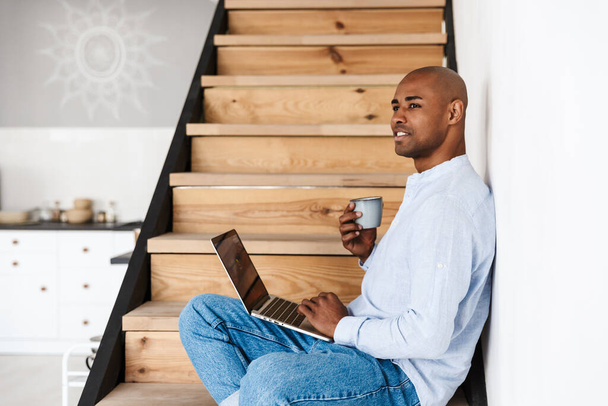 Photo of smiling african american man using laptop and drinking coffee while sitting on wooden stair at home - Φωτογραφία, εικόνα