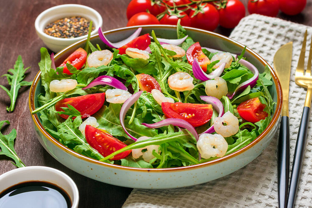 Mediterranean diet menu concept Healthy salad of fresh vegetables - tomatoes, arugula, purple onions in plate, sesame seed, soy sauce, knife, fork, napkin on wooden table Vegan food Flat lay Top view. - Photo, Image