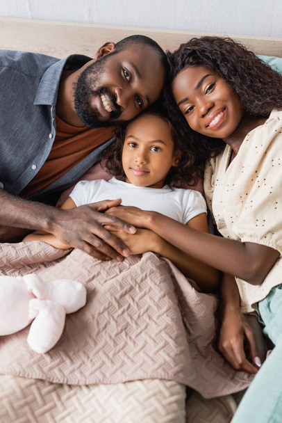alto ángulo vista de afroamericano marido y esposa tocando manos de hija acostada en la cama - Foto, Imagen