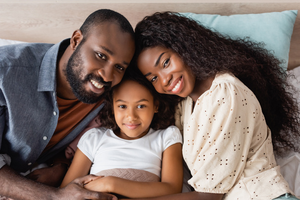 high angle view of african american couple looking at camera near daughter lying in bed  - Фото, зображення